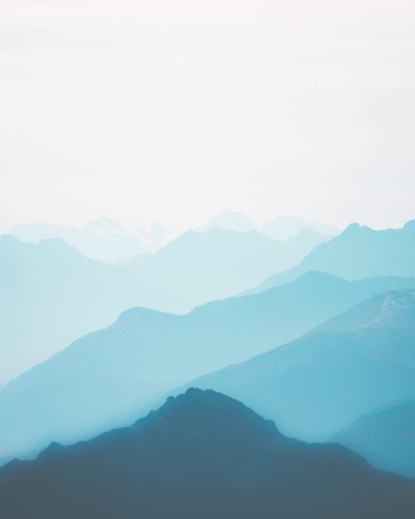 Diverse bergtoppen in verschillende tinten blauw tegen een lichte lucht.