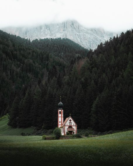 Een kleine kerk omgeven door bomen en bergen in een mistig landschap.